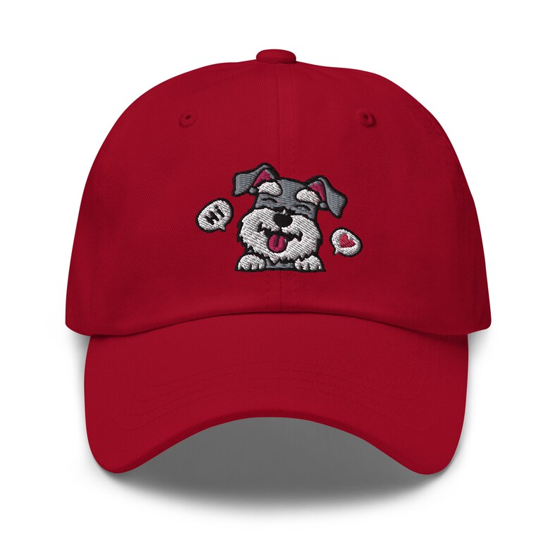 K&ouml;nnte beinhalten: Rote Baseballkappe mit einem gestickten Cartoon-Bild eines grau-wei&szlig;en Zwergschnauzers mit herausgestreckter rosa Zunge. Der Hund tr&auml;gt ein rotes Herz und hat eine Sprechblase mit der Aufschrift "Hallo".