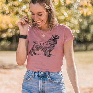May include: A dusty rose t-shirt featuring a black illustration of a Cocker Spaniel dog. The dog is depicted in a detailed, side-profile pose. The shirt has a classic crew neck and short sleeves, paired with blue denim shorts.