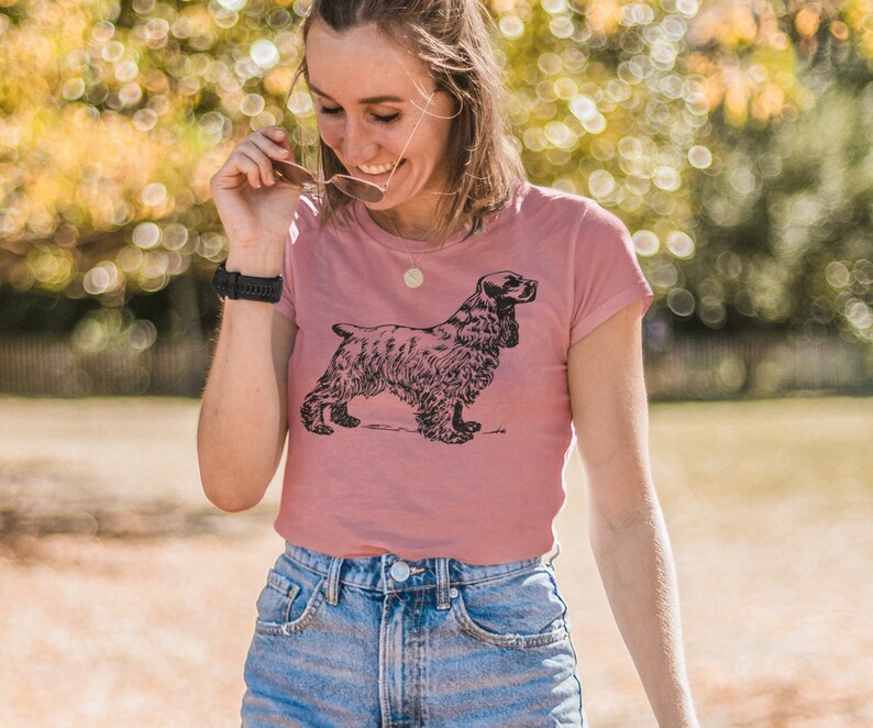May include: A dusty rose t-shirt featuring a black illustration of a Cocker Spaniel dog. The dog is depicted in a detailed, side-profile pose. The shirt has a classic crew neck and short sleeves, paired with blue denim shorts.