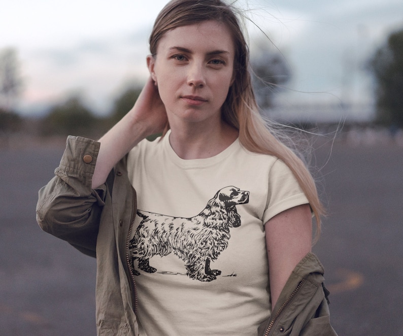 May include: Cream-colored t-shirt featuring a detailed black and white illustration of a Cocker Spaniel dog. The dog is depicted in a standing pose, showcasing its distinctive features. The shirt is worn by a person.