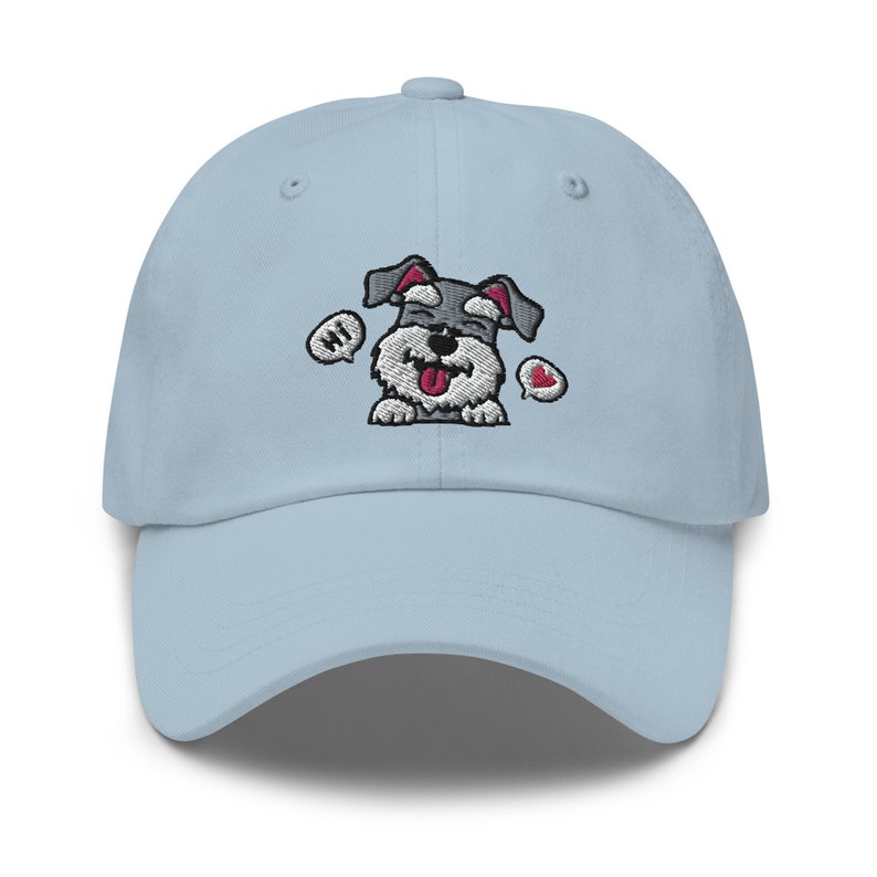 K&ouml;nnte beinhalten: Eine hellblaue Baseballkappe mit einem gestickten Cartoon-Bild eines grau-wei&szlig;en Zwergschnauzers mit herausgestreckter roter Zunge. Der Hund sagt "Hallo" in einer Sprechblase und hat ein rotes Herz &uuml;ber seinem Kopf.