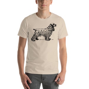 May include: A beige t-shirt featuring a black illustration of a Cocker Spaniel dog. The dog is depicted in a standing profile, showcasing its distinctive features. The shirt is a casual wear item.