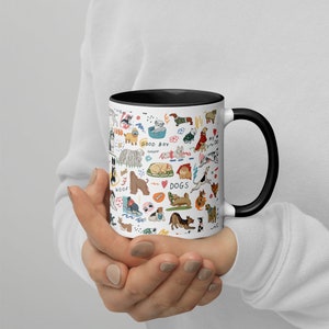 Op de afbeelding: Een witte keramische koffiemok met een zwarte rand en handvat. De mok heeft een kleurrijke illustratie van verschillende hondenrassen, waaronder een teckel, een poedel, een golden retriever en een bulldog. De illustratie bevat ook de woorden "Good Boy", "Woof" en "Dogs".