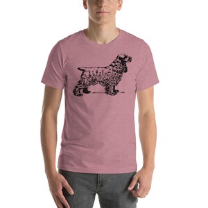 May include: A short-sleeved, mauve t-shirt featuring a black illustration of a Cocker Spaniel dog. The dog is depicted in a side profile, showcasing its distinctive long ears and fluffy fur. The shirt is made of a soft material.