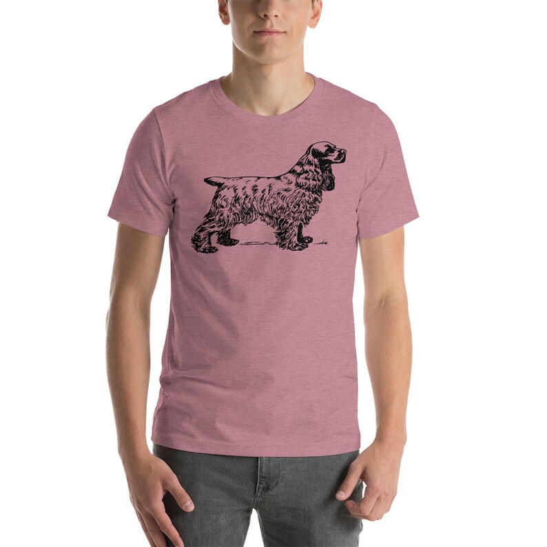 May include: A short-sleeved, mauve t-shirt featuring a black illustration of a Cocker Spaniel dog. The dog is depicted in a side profile, showcasing its distinctive long ears and fluffy fur. The shirt is made of a soft material.