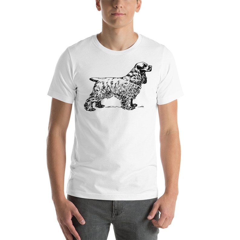 May include: White t-shirt featuring a black and white illustration of a Cocker Spaniel dog. The dog is depicted in a standing pose, showcasing its distinctive features. The shirt is a classic crew neck style.