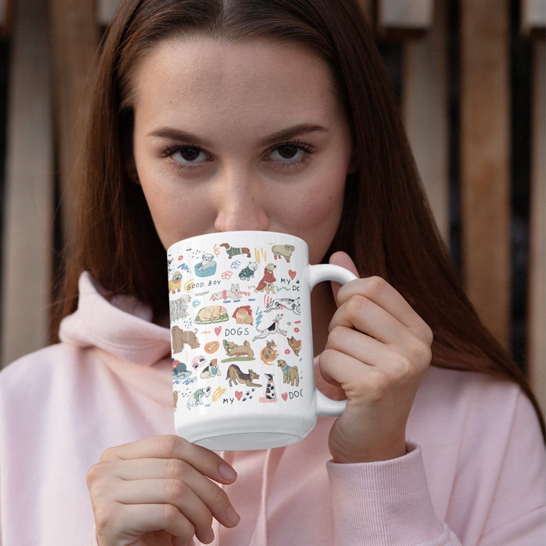 May include: A white ceramic mug with a colorful illustration of dogs and the words "Good Boy", "My Dog", and "Dogs" on it. The mug is being held by a person.