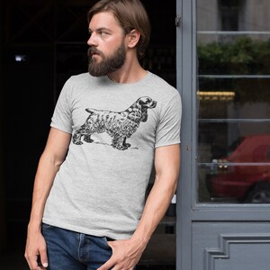 May include: A light gray t-shirt featuring a detailed black and white illustration of a Cocker Spaniel dog. The dog is standing in profile, showcasing its distinctive features. The shirt is a classic crew neck style.