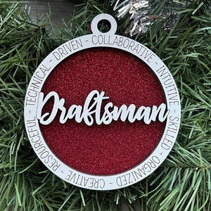May include: A white wooden ornament with a red glitter background. The ornament has the word "Draftsman" written in white cursive lettering. The words "COLLABORATIVE - INTUITIVE - SKILLED - ORGANIZED - CREATIVE - RESOURCEFUL - TECHNICAL - DRIVEN" are written around the edge of the ornament.