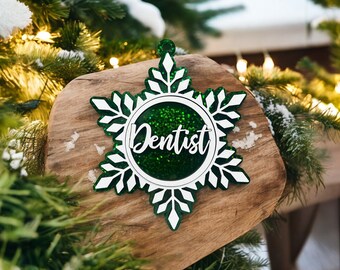 May include: A snowflake-shaped Christmas ornament with the word "Dentist" in white script. The ornament has a green glitter center and white and green snowflake edges. It is displayed on a wooden surface with a snowy background.