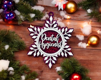 May include: A snowflake-shaped Christmas ornament with the words "Dental Hygienist" in a circular design. The ornament is purple with white accents and glitter. The background includes Christmas decorations such as ornaments, pine branches, and snow.