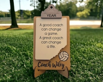 May include: A tan wooden clipboard-style sign featuring the text 'YEAR' at the top, a volleyball graphic, and the name 'Coach Wiley'. The center displays the quote: 'A good coach can change a game. A great coach can change a life.'