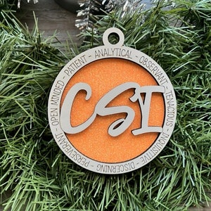 May include: A round Christmas ornament with the letters "CSI" in the center, surrounded by words like "Analytical" and "Patient." The ornament has an orange center and a silver-colored border. It is displayed on a bed of green garland.
