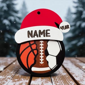 May include: A Christmas ornament featuring a basketball, football, and soccer ball design. The ornament has a red Santa hat with the word "NAME" and a small circle that says "YEAR". The ornament is on a wooden surface.