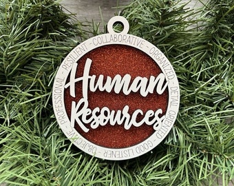 May include: A circular wooden ornament with the words "Human Resources" in a cursive font, set against a red glitter background. The outer ring features words like "Professional" and "Collaborative". The ornament is displayed on green pine needles.