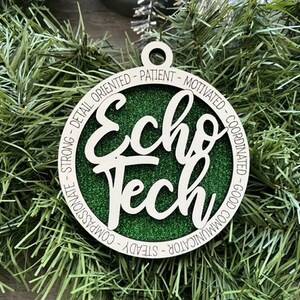 May include: A round, wooden Christmas ornament with the words "Echo Tech" in a cursive font, layered over a green glitter background. The outer ring features words like "compassionate" and "motivated". The ornament is hanging on a bed of green pine needles.