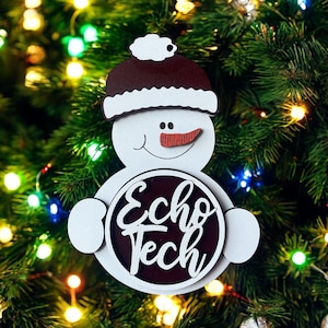 May include: A white snowman Christmas ornament with a brown hat and a red carrot nose. The ornament has the text "Echo Tech" in a circular design. The ornament is hanging on a Christmas tree with colorful lights.