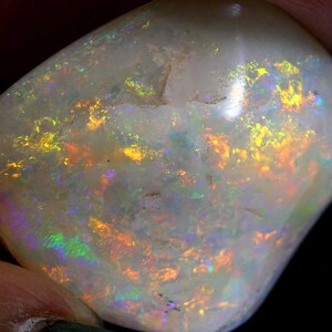 Może przedstawiać: Wypolerowany, owalny kamień szlachetny z opalu. Kamień ma mlecznobiały kolor bazowy z żywymi błyskami ż&oacute;łci, pomarańczy i zieleni, tworząc efekt iryzacji. Opal trzymany jest między palcami, na ciemnym tle.