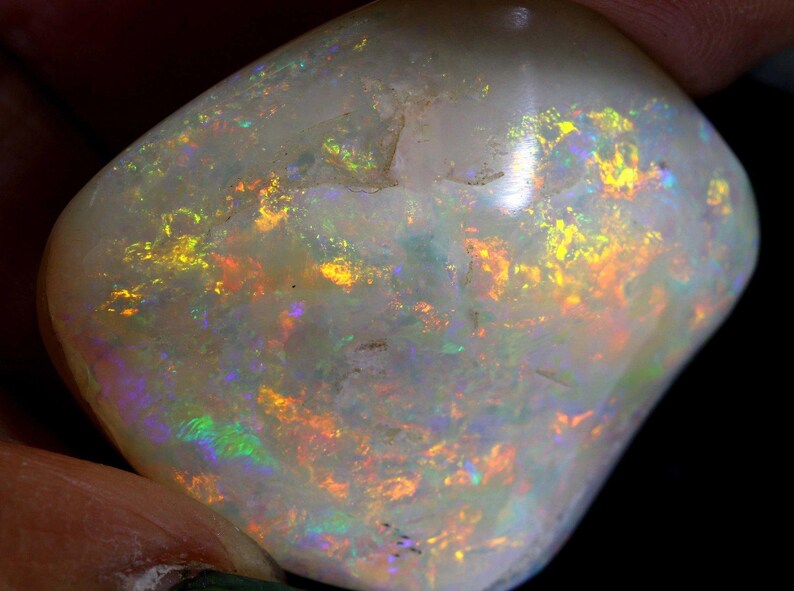 Może przedstawiać: Wypolerowany, owalny kamień szlachetny z opalu. Kamień ma mlecznobiały kolor bazowy z żywymi błyskami ż&oacute;łci, pomarańczy i zieleni, tworząc efekt iryzacji. Opal trzymany jest między palcami, na ciemnym tle.