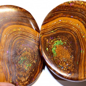 Könnte beinhalten: Zwei runde, braune und goldene Opal-Cabochons mit irisierenden grünen und violetten Blitzen. Die Opale haben ein wirbelndes, geschichtetes Muster.