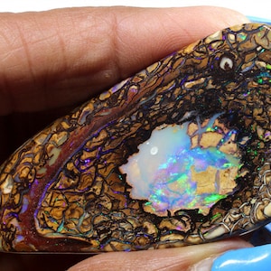 Könnte beinhalten: Ein großes, unregelmäßig geformtes Stück Opal mit einer braunen, schwarzen und weißen Matrix. Der Opal hat einen leuchtenden, irisierenden Farbspiel in Blau, Grün und Violett.