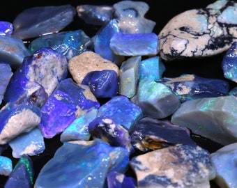 Puede incluir: Una colección de piedras preciosas de ópalo natural en bruto en varios tonos de azul y verde. Los ópalos tienen un brillo iridiscente y se muestran sobre un fondo negro.