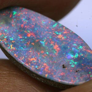 May include: Oval-shaped opal gemstone with a dark base and a vibrant play of color. The stone displays flashes of red, green, and blue throughout its surface. The opal is set against a neutral background, highlighting its iridescent qualities.