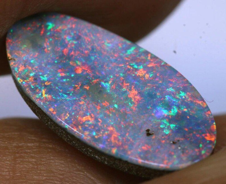 May include: Oval-shaped opal gemstone with a dark base and a vibrant play of color. The stone displays flashes of red, green, and blue throughout its surface. The opal is set against a neutral background, highlighting its iridescent qualities.