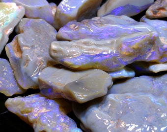 Puede incluir: Una colección de piedras de ópalo en bruto, grises, con matices iridiscentes azules y morados. Las piedras tienen una forma irregular y una superficie lisa y pulida.