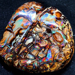 Op de afbeelding: Een grote, hartvormige opaal met een bruine, witte en zwarte matrix. De opaal vertoont een levendig, iriserend kleurenspel, waaronder blauw, groen, rood en oranje.