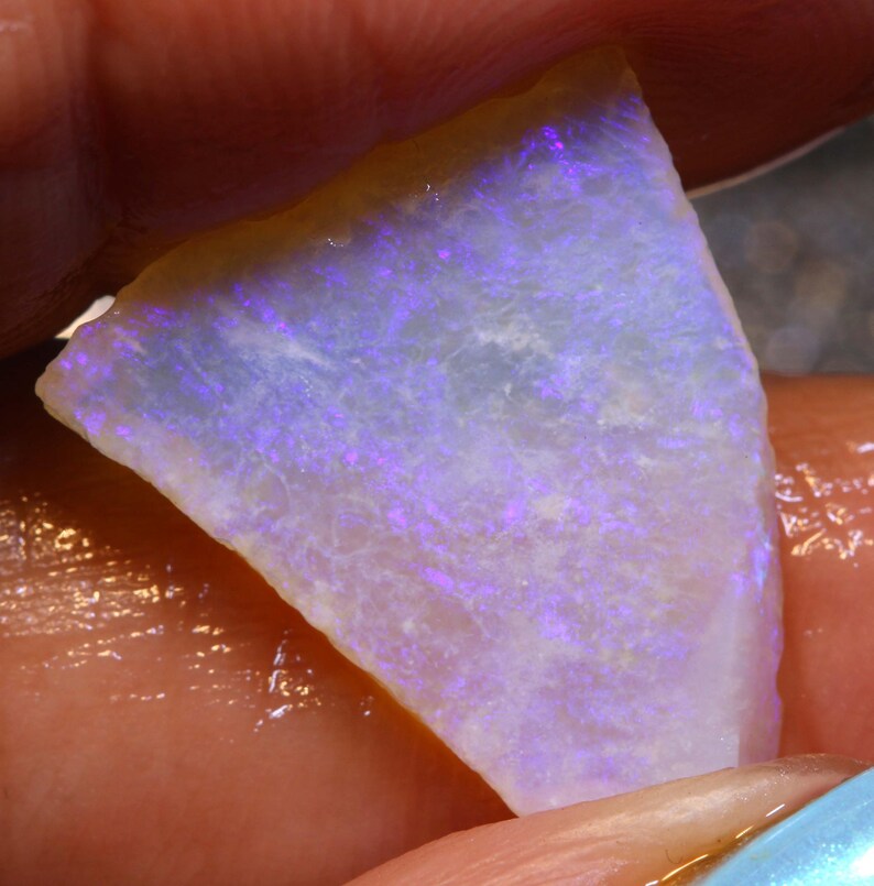 May include: A triangular-shaped opal gemstone with a milky white base and vibrant flashes of purple and blue. The stone has a rough, natural texture and a thin, golden-brown edge. The opal is held in a hand, showcasing its iridescent play of color.