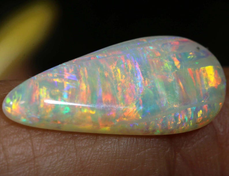 Pu&ograve; includere: Una gemma di opale a forma di goccia, con una base bianco latte. L'opale mostra vivaci lampi di colore, tra cui rosso, arancione, giallo, verde e blu, creando un effetto iridescente. La gemma &egrave; liscia e lucidata.