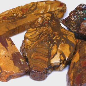 Puede incluir: Una colecci&oacute;n de piedras preciosas en bruto, sin cortar, en tonos marr&oacute;n, beige y marr&oacute;n rojizo. Las piedras tienen patrones y texturas &uacute;nicos, algunos con apariencia veteada. Las piedras preciosas se exhiben sobre una superficie blanca.