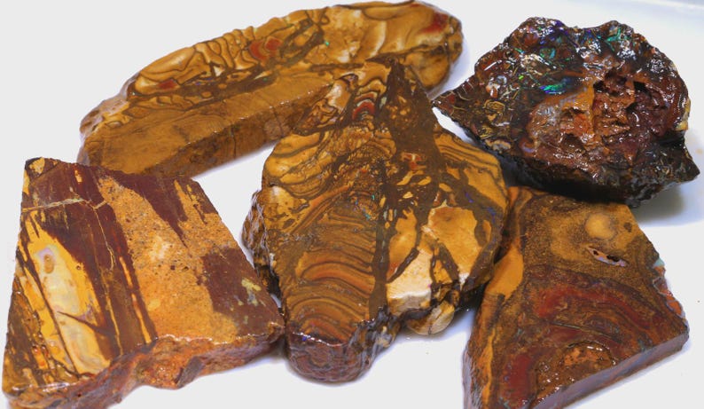 Puede incluir: Una colecci&oacute;n de piedras preciosas en bruto, sin cortar, en tonos marr&oacute;n, beige y marr&oacute;n rojizo. Las piedras tienen patrones y texturas &uacute;nicos, algunos con apariencia veteada. Las piedras preciosas se exhiben sobre una superficie blanca.