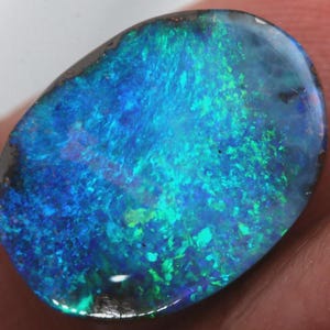 Peut inclure: Une pierre précieuse opale de forme ovale, affichant un jeu de couleurs éclatant. La pierre présente un mélange de bleus et de verts, avec une bordure sombre. La surface de l'opale est polie.