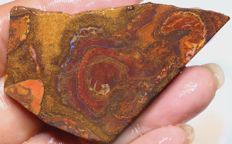 Puede incluir: Una gema triangular pulida con una vibrante exhibici&oacute;n de colores. La piedra presenta patrones arremolinados de rojo intenso, naranja y dorado, con toques de azul. La textura general parece &aacute;spera y natural, mostrando la belleza &uacute;nica del mineral.