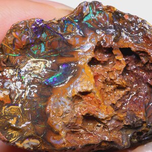 Puede incluir: Una piedra preciosa de &oacute;palo en bruto, sin cortar, con una variedad de colores. El &oacute;palo muestra una mezcla de tonos marrones, naranjas y negros, con destellos iridiscentes de azul, verde y p&uacute;rpura. La piedra tiene una superficie texturizada e irregular.