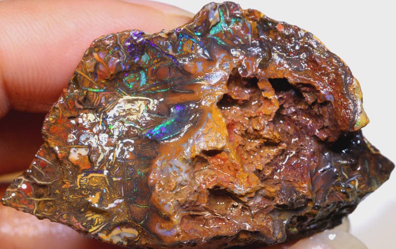 Puede incluir: Una piedra preciosa de &oacute;palo en bruto, sin cortar, con una variedad de colores. El &oacute;palo muestra una mezcla de tonos marrones, naranjas y negros, con destellos iridiscentes de azul, verde y p&uacute;rpura. La piedra tiene una superficie texturizada e irregular.