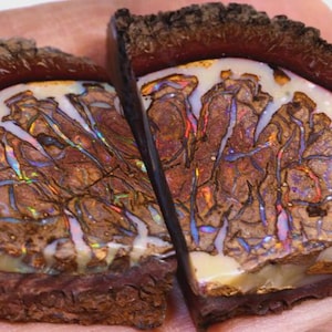 Op de afbeelding: Twee helften van een gepolijste opaal edelsteen. De opaal heeft een crèmewitte basis met ingewikkelde bruine en iriserende patronen. De buitenste randen zijn donkerbruin en getextureerd, waardoor een natuurlijke lijst voor de kleurrijke steen ontstaat.