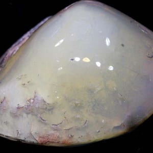 May include: A polished, rounded gemstone in a pale yellow-green shade. The stone is translucent, with delicate colour and texture variations. The surface reflects light, creating highlights. The gemstone is set against a black background.