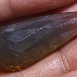 May include: A teardrop-shaped, faceted gemstone pendant with a hole at the top for stringing. The gemstone is a mix of gray, brown, and green with a slight iridescent sheen.