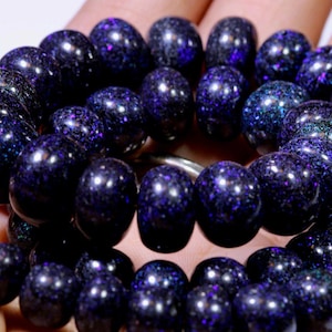 Può includere: Un braccialetto di perline blu scuro con macchie iridescenti. Le perline rotonde sono infilate su un cordoncino bianco. Il braccialetto è tenuto in una mano, mostrando il colore e la consistenza delle perline. Le perline hanno una finitura lucida e lucente.