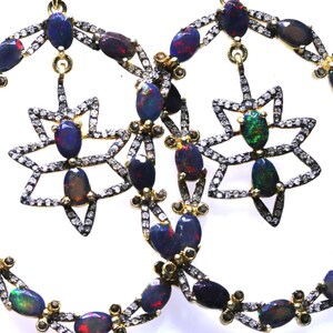 May include: A pair of hoop earrings featuring oval-shaped, dark blue opals with red and green flashes. The earrings have a star-shaped pendant and are adorned with small diamonds and gold accents. The opals are set in a silver-colored metal.