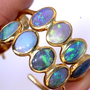 May include: A gold hoop earring with a series of oval opals. The opals display blues, greens, and purples, with an iridescent effect. The earring has a continuous, flowing shape. The opals are set in gold. The earring is a piece of jewellery.