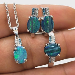 Op de afbeelding: Een sieradenset met een zilveren ketting, ring en oorbellen. Elk stuk toont een ovale opaal met blauwe en groene tinten, geaccentueerd met kleine, heldere en blauwe edelstenen. De opaalhanger hangt aan een fijne ketting.