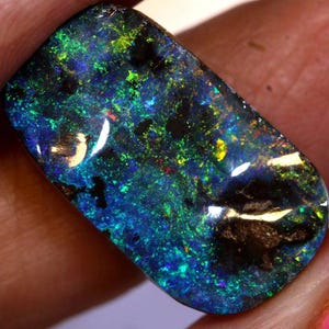 May include: A polished black opal gemstone with vibrant flashes of blue, green, and yellow. The rectangular stone displays a play of color, with dark areas contrasting the bright hues. The opal is held in a hand, showcasing its natural beauty.
