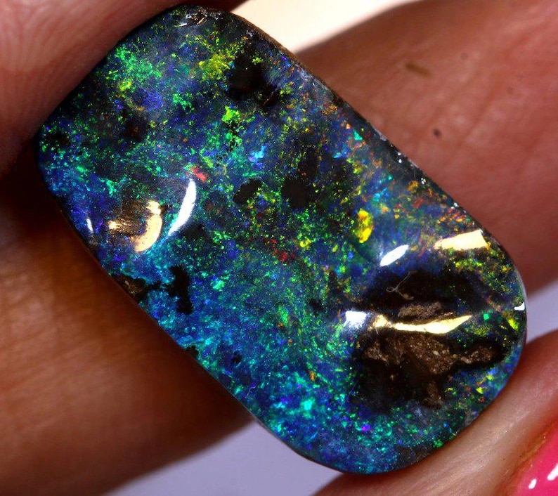 May include: A polished black opal gemstone with vibrant flashes of blue, green, and yellow. The rectangular stone displays a play of color, with dark areas contrasting the bright hues. The opal is held in a hand, showcasing its natural beauty.