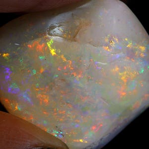 Może przedstawiać: Wypolerowany, opalizujący kamień szlachetny z opalu. Kamień prezentuje mlecznobiałą barwę podstawową z błyskami żywych kolor&oacute;w, w tym czerwonego, pomarańczowego, ż&oacute;łtego, zielonego i niebieskiego. Opal jest trzymany przez palce osoby na czarnym tle.