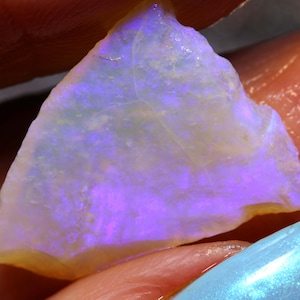 May include: A close-up of a raw opal gemstone. The opal has a milky white base color with flashes of purple and blue iridescence. The edges of the stone have a yellowish hue.