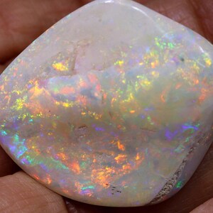 Może przedstawiać: Wypolerowany, biały opal z żywymi, iryzującymi błyskami koloru. Kamień szlachetny prezentuje grę światła z odcieniami pomarańczowego, ż&oacute;łtego, zielonego i niebieskiego. Opal jest gładki i zaokrąglony, ukazując swoje naturalne piękno.